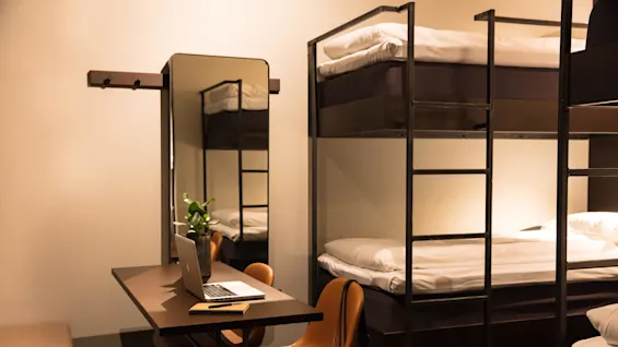 Laptop rests on a table beside a small plant and notebook, surrounded by orange chairs. Nearby, bunk beds with white linens are visible, in a warmly lit room.