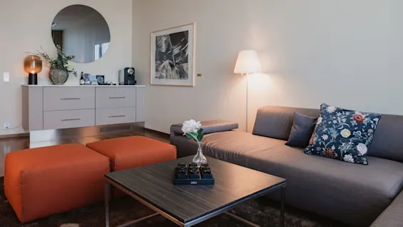 A gray sofa with floral cushions sits in a modern living room, accompanied by a table with a flower vase. Nearby, two orange poufs stand beside a cabinet with decor and a mirror.