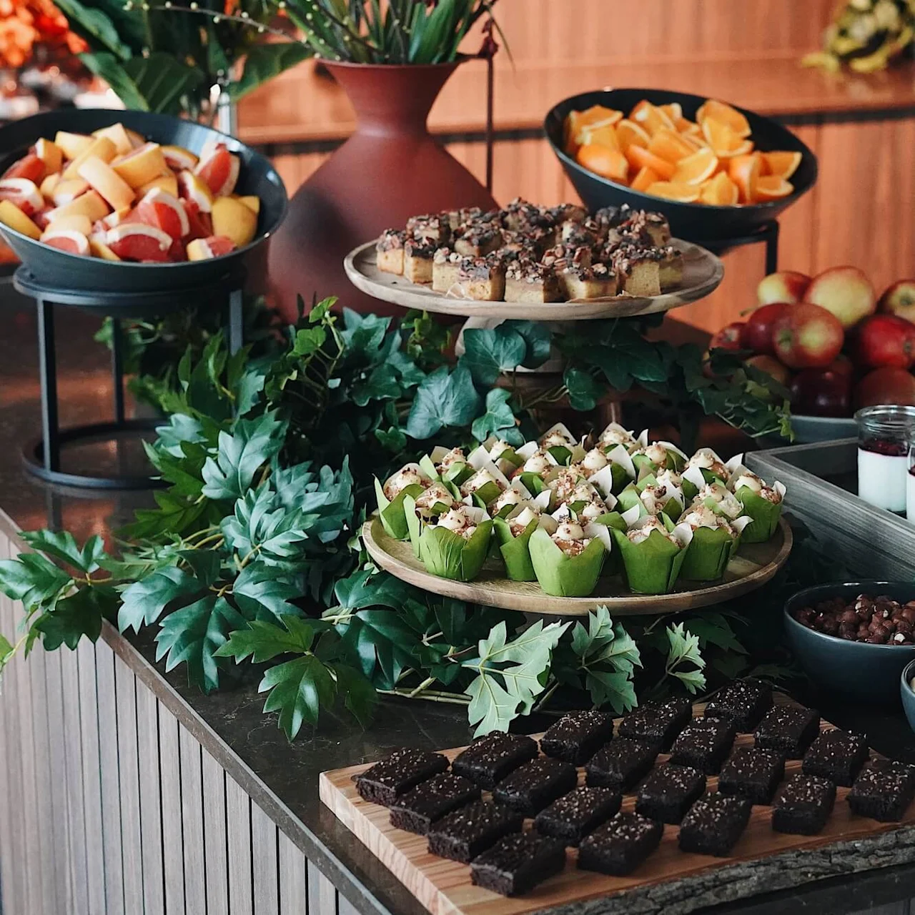Olika desserter och frukter visas elegant på en buffé med gröna accenter. Olika bakverk, fruktskivor och burkar med yoghurt skapar en färgstark, inbjudande uppsättning.