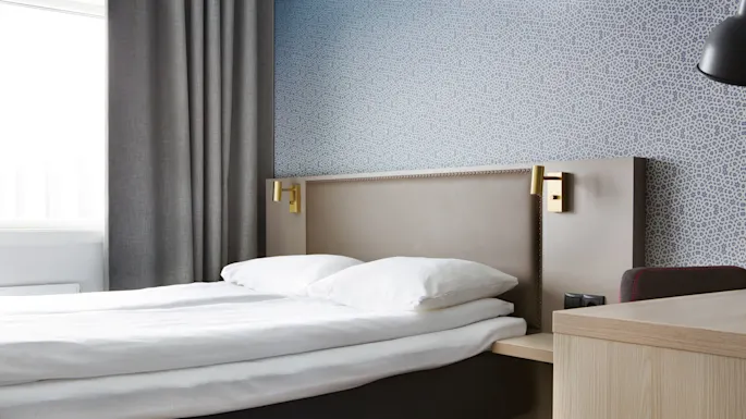 A neatly made bed with white bedding in a room with patterned wallpaper and a window with grey curtains.