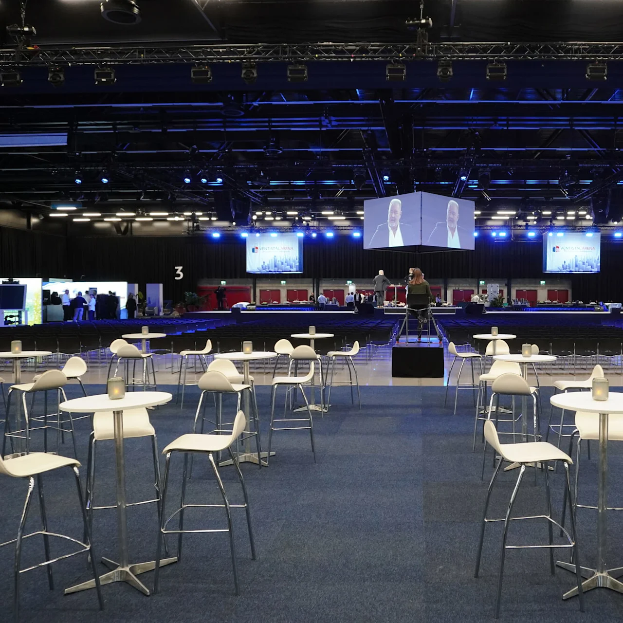 Bord och stolar står uppställda i en tom konferenshall med stora skärmar som visar ett evenemang. I taket finns rigg och belysning, och flera bås syns.