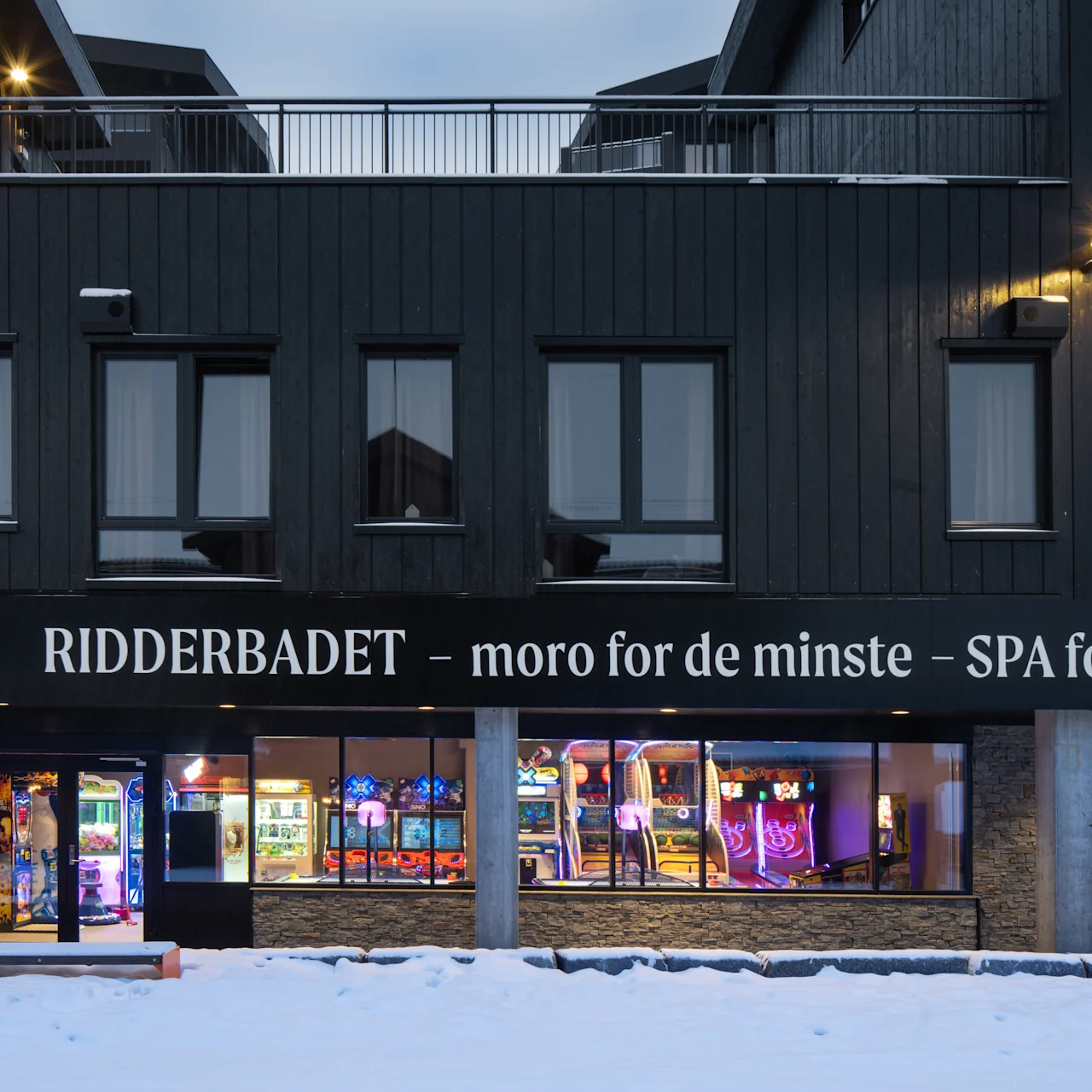 Mørk bygningsfacade med teksten "RIDDERBADET – moro for de minste – SPA for de voksne." Lyse spillemaskiner er synlige indeni gennem store vinduer. Beliggende i et sneklædt landskab.