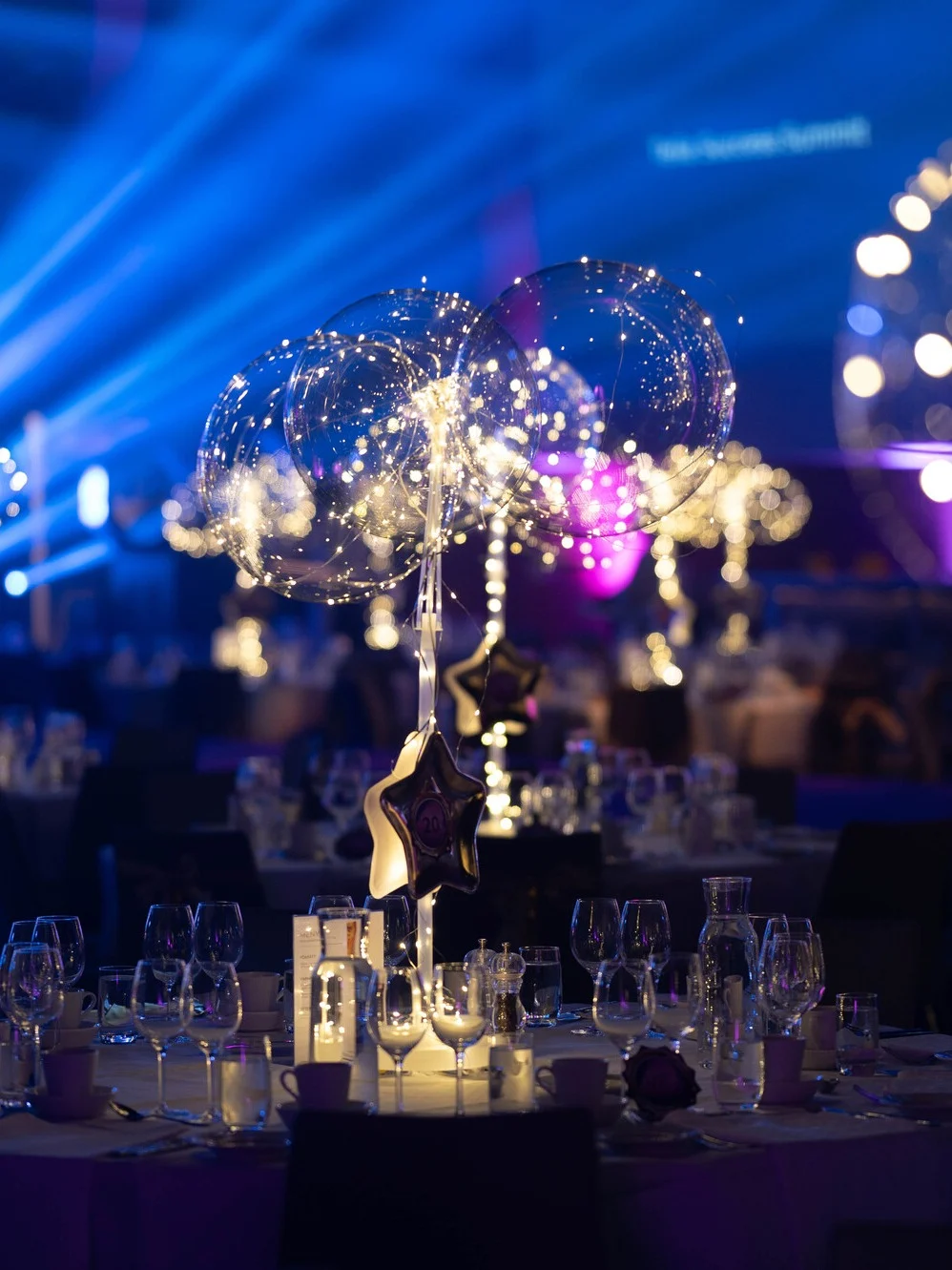 Bord dekorerade med upplysta mittstycken med glasbubblor och feerljus; i en svagt upplyst bankettsal med blå och lila omgivningsljus. Text på skärm: "Next Awards Summit."