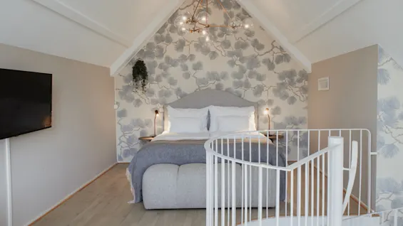 Et hyggeligt soveværelse med en stor seng, mønstret tapet, et fjernsyn og en hvid vindeltrappe, der fører op.