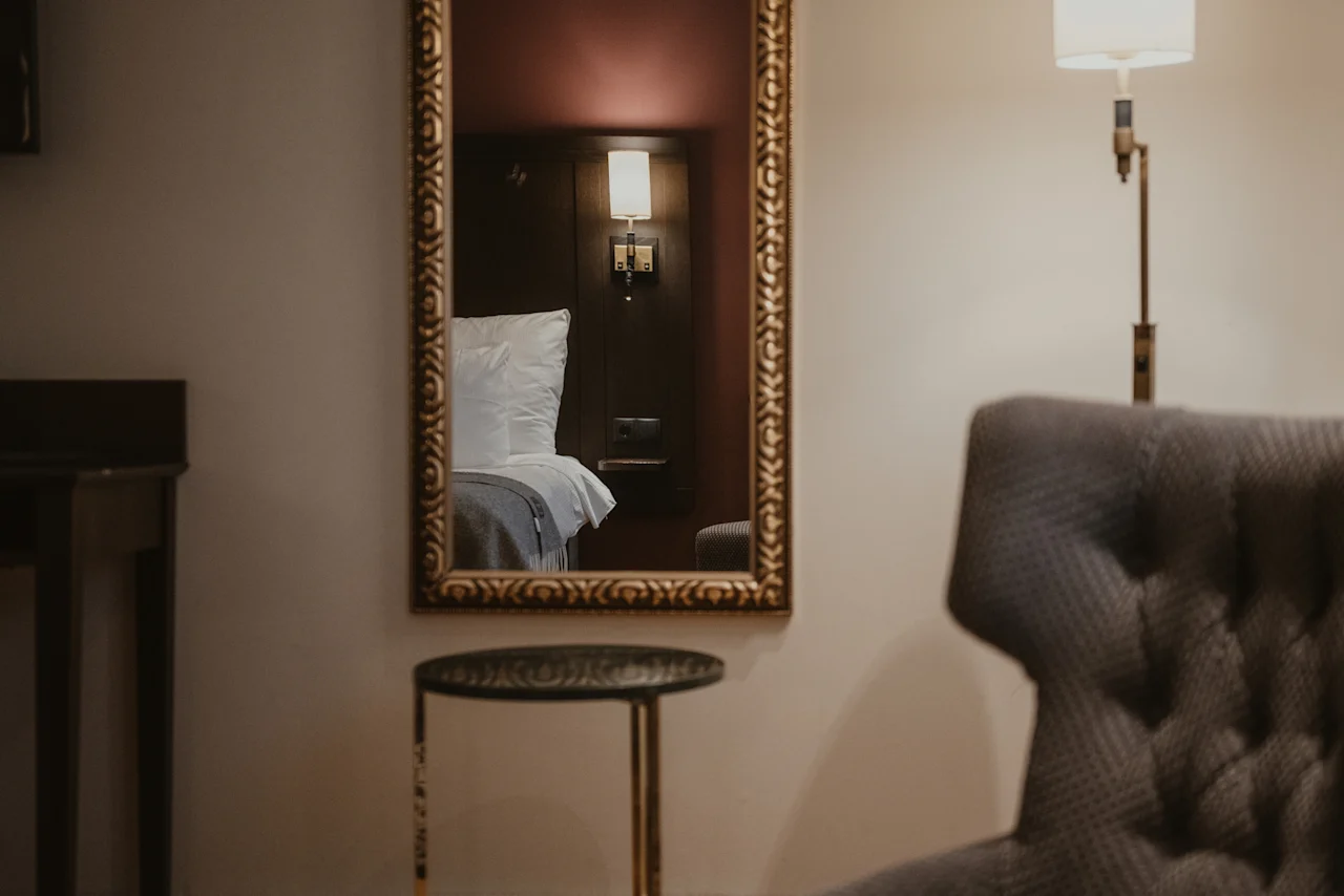 A cozy hotel room with a mirror reflecting a comfortable bed, an armchair, and a small table. add Home Hotel Kung Oscar