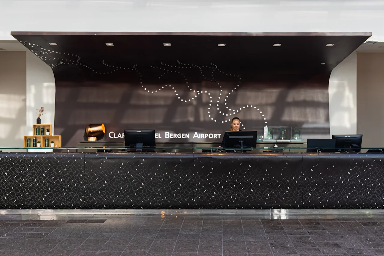 En lang resepsjonsdisk med datamaskiner og en ansatt står i en moderne lobby. Bak en svart vegg er prikkete kunst og teksten "CLARION HOTEL BERGEN AIRPORT."