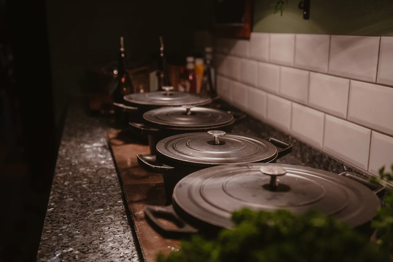 Rivi tummia, kannellisia keittoastioita tiskillä keittiöympäristössä, taustalla valkoiset metrolaatat.