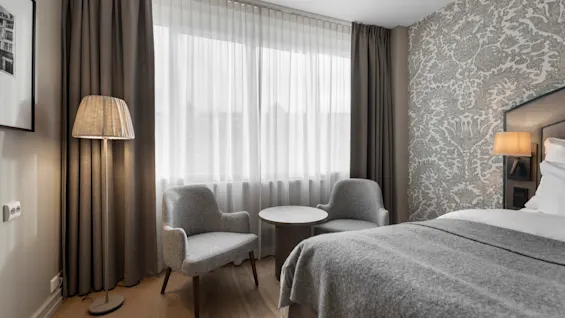 A cozy hotel room with a bed, two armchairs, and a small table by the window, creating a comfortable space for relaxation.