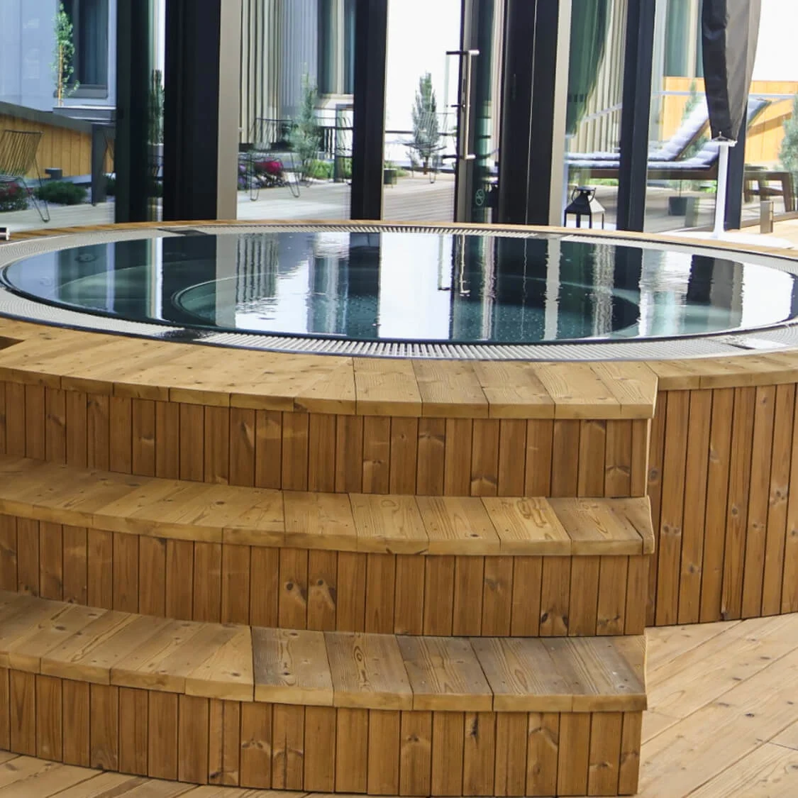 A round wooden hot tub with steps is set on a deck, surrounded by glass walls, reflecting a modern outdoor patio with plants and chairs.