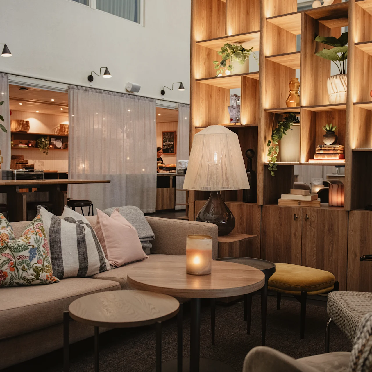 En mysig lounge har beige soffor med färgglada kuddar, ett runt träbord med ett tänt ljus och en hög hylla med böcker och växter i ett svagt upplyst rum.