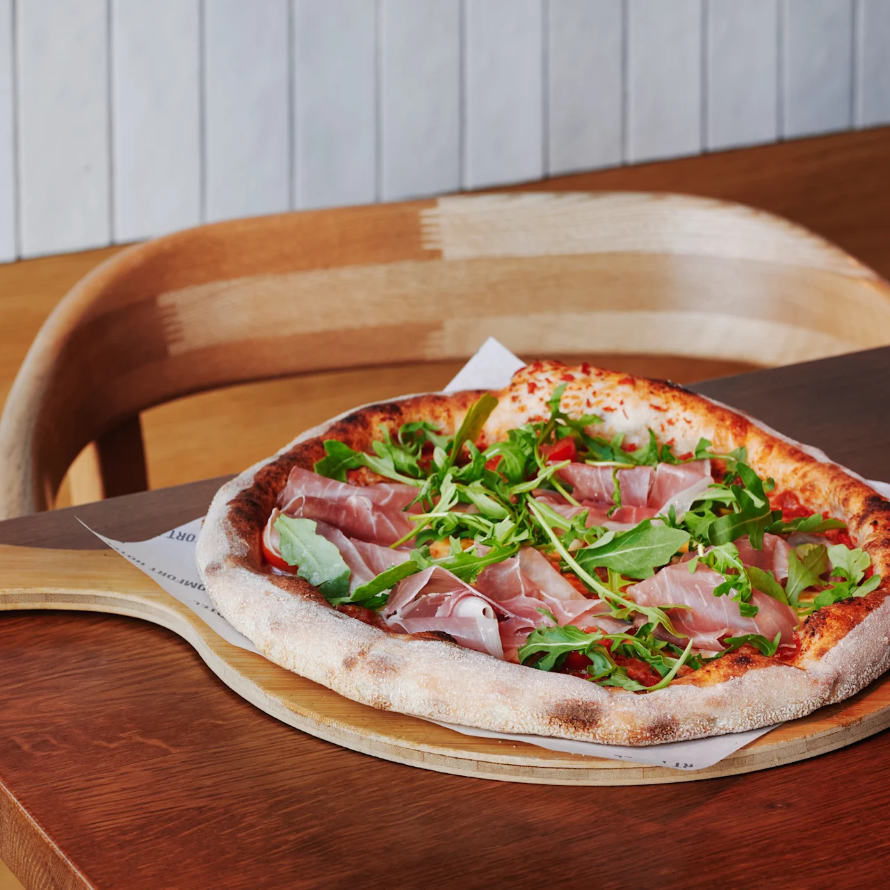 Wood-fired pizza topped with prosciutto and arugula sits on a wooden paddle, placed on a wooden table in a cozy dining setting with a wooden chair in the background.
