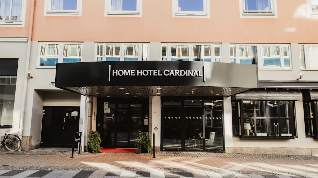 Kutsuva hotellin sisäänkäynti, jossa kyltti HOME HOTEL CARDINAL. Punainen matto ja ruukkukasvit koristavat sisäänkäyntiä.