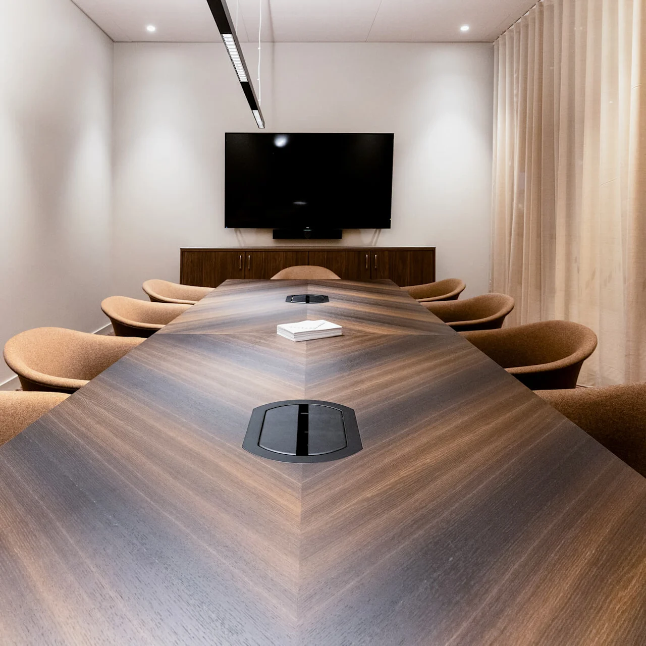 A long, polished wooden conference table surrounded by brown chairs in a meeting room, facing a large wall-mounted screen with beige curtains along the side.