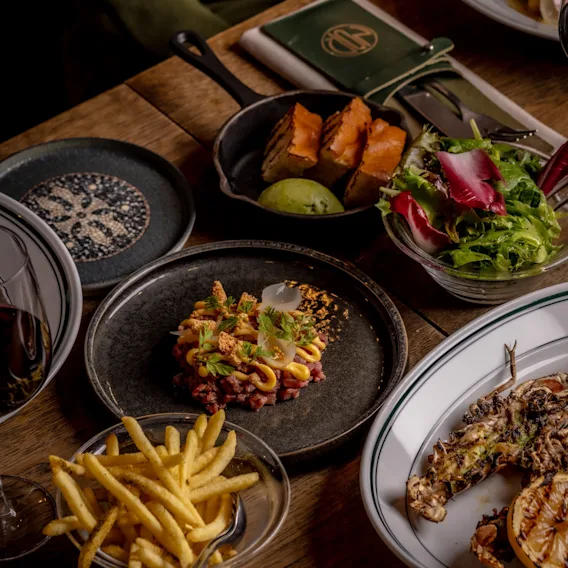 A table laden with various delicious dishes, including steak tartare, grilled seafood, fries, and salad, ready for a meal.