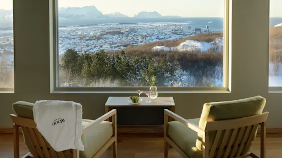 Et hyggeligt værelse med to stole og et bord, der byder på en fantastisk vinterudsigt over et sneklædt landskab, bjerge og en by. WOOD