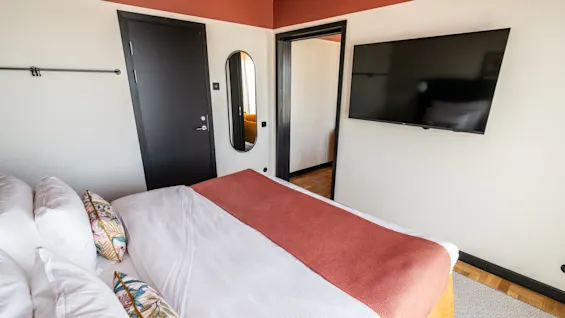 A bed with vibrant pillows and a red blanket sits in a sleek hotel room. A black door, oval mirror, and mounted TV adorn the walls.