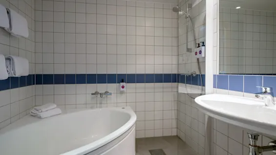 Badekar med hvide håndklæder læner mod flisebelagte vægge under en blå stribe. Ved siden af står en bruser med monterede dispensere ved siden af en slank vask.