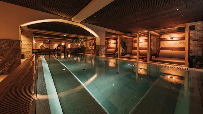 Et rektangulært innendørsbasseng med dempet belysning, omgitt av loungemøbler og trebjelker, gir en varm og innbydende atmosfære for avslapning.