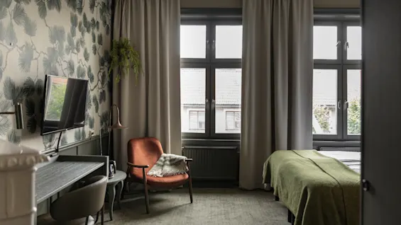 Viihtyisä hotellihuone, jossa on sänky, työpöytä ja mukava tuoli, suuret ikkunat ja luontoaiheinen tapetti.