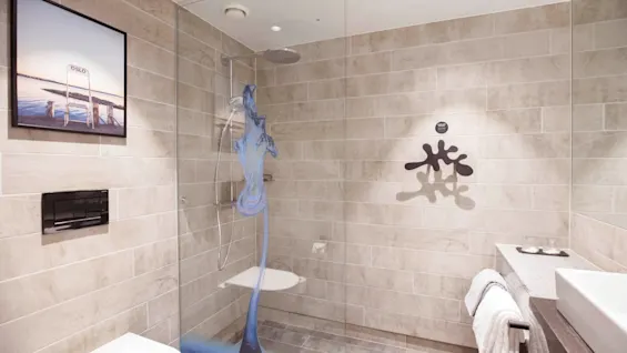A modern bathroom features a glass-enclosed shower with a blue water sculpture, beige tiles, a wall-mounted toilet, and a picture frame. Towels hang beside a rectangular sink.