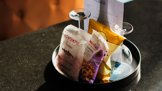 En minibar bakke med snacks som KONFEKTIR og NUTISAL, en lille flaske og to glas, klar til at nydes.