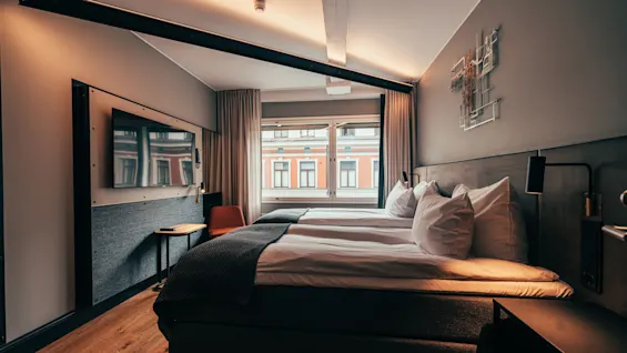 A modern hotel room with two comfortable beds, a wall-mounted TV, and a window offering a city view.