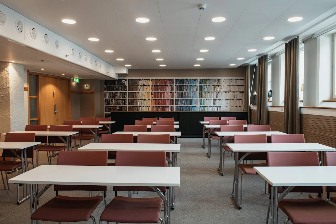 A bright conference room at Home Hotel Uppsala with rows of tables and chairs, and a wall decorated with a bookshelf mural.