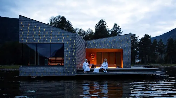English: A modern building with angular architecture floats on water. Two people sit outside wrapped in white robes. It's surrounded by trees and mountains under a cloudy sky.