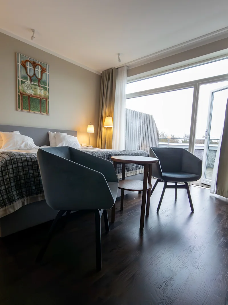 A cozy hotel room with a comfortable bed, two armchairs, and a large window offering a bright view.