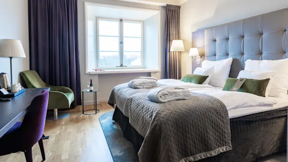 A neatly arranged hotel room features a large bed with white linens, a chair next to a desk, a floor lamp, and a window with curtains. A small table holds a wine bottle and glasses.