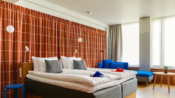 En pænt redt dobbeltseng med gråt og hvidt sengetøj står i et moderne soveværelse. Lyse orange gardiner og en blå sofa skaber en livlig, hyggelig atmosfære.