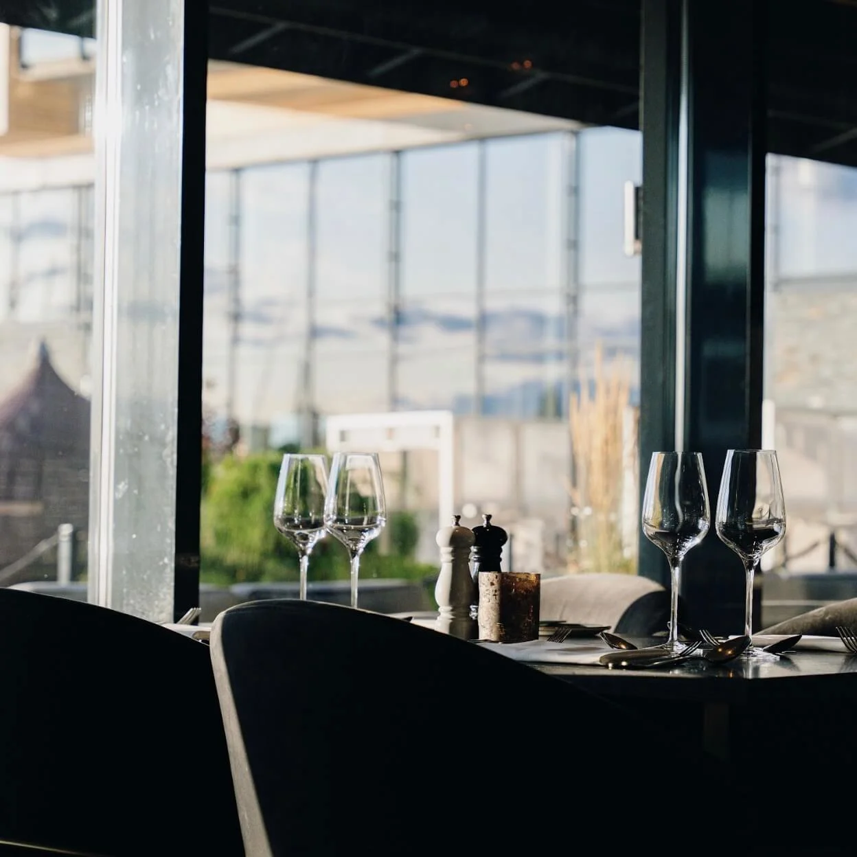 Et dekket bord med tomme vinglass er plassert innendørs nær store vinduer, med utsikt over en solfylt utendørs terrasse.