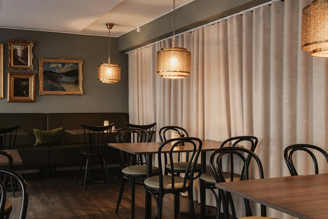 En inbjudande matplats med träbord, svarta stolar, en bås och varma, dekorativa taklampor. Konst hänger på väggen.