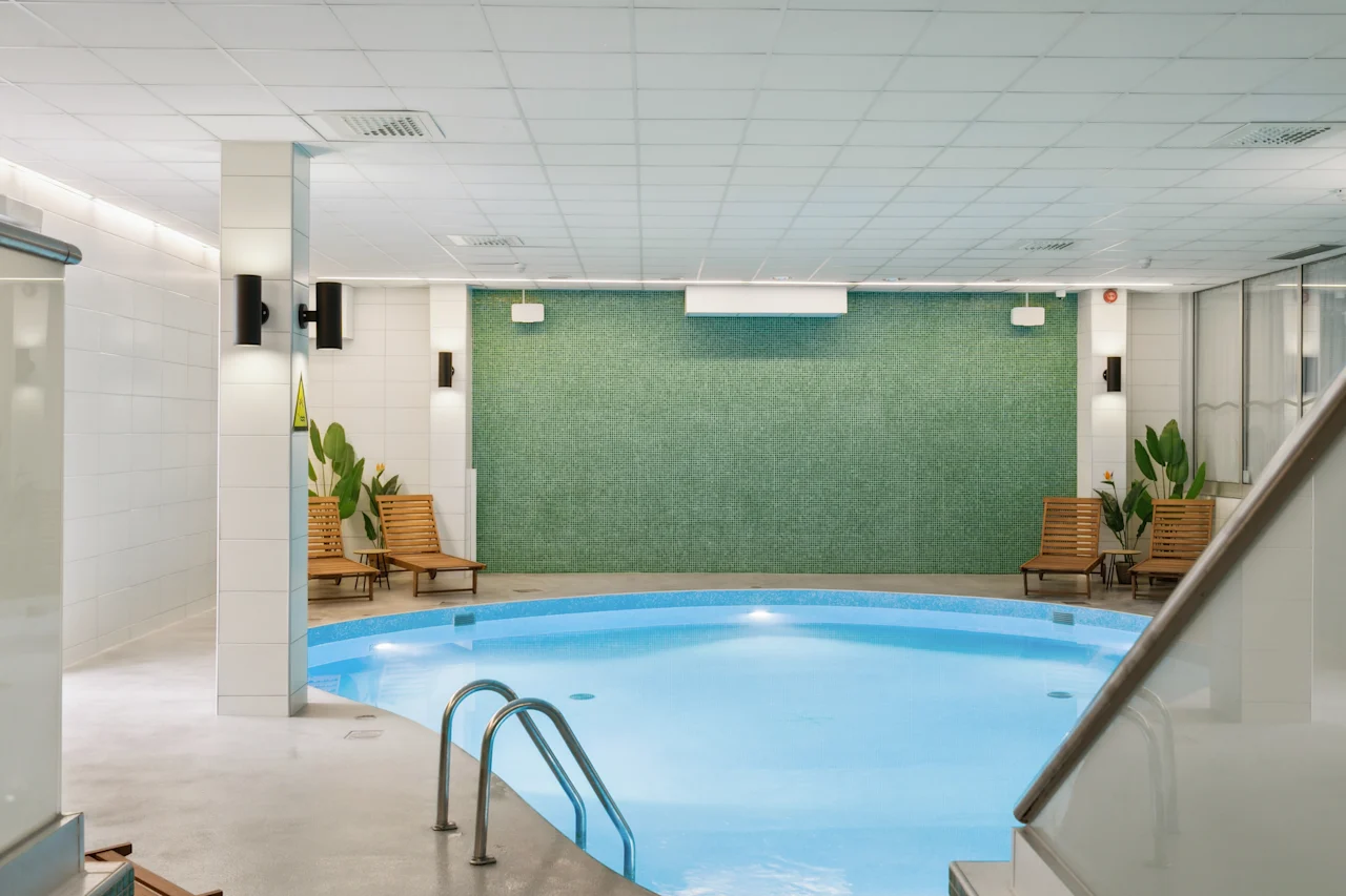 En lugn inomhuspool vilar stillsamt, omgiven av solstolar och krukväxter mot vitkaklade väggar. Den fridfulla miljön är mjukt upplyst under ett gallerformat tak.
