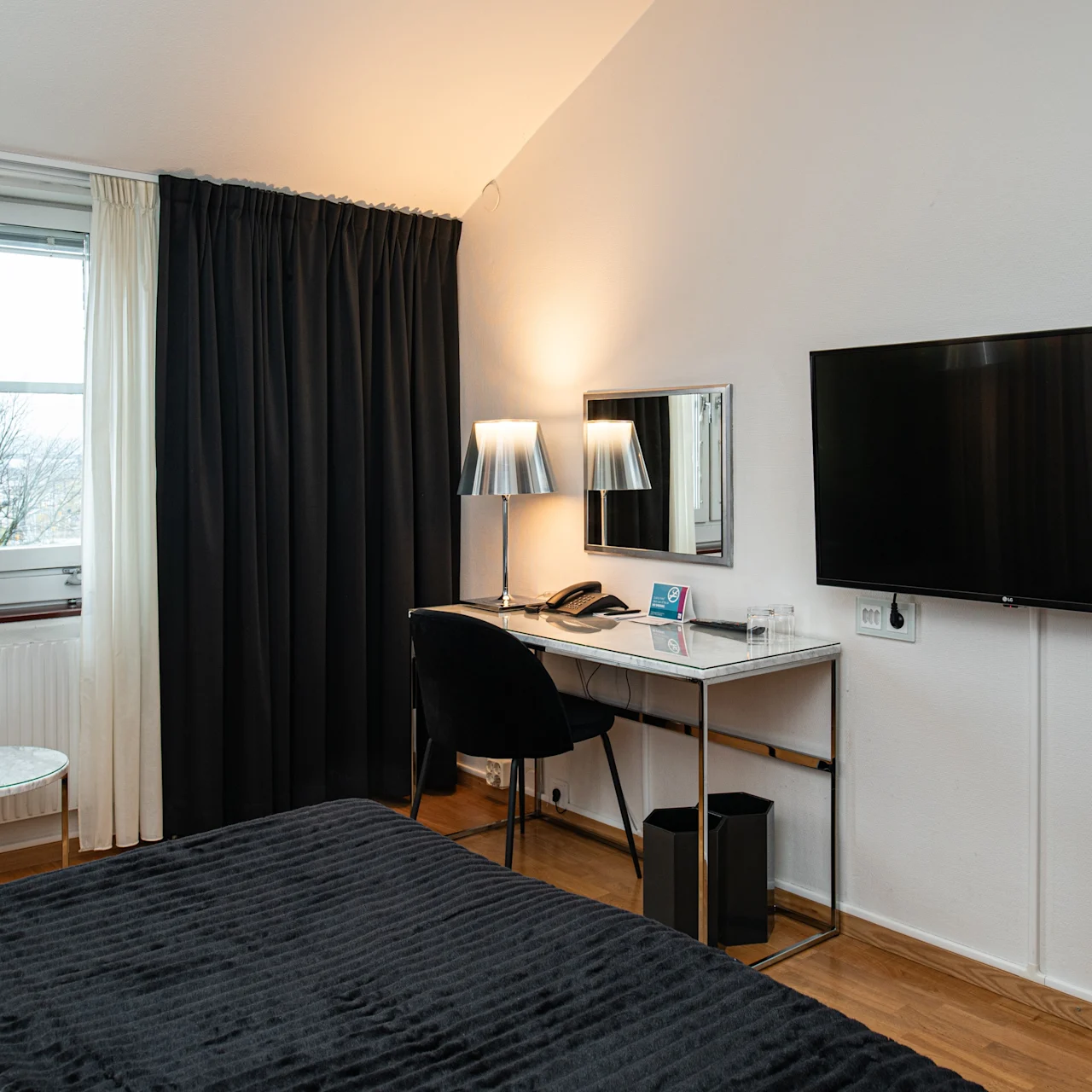 Et pænt indrettet hotelværelse med en seng, et skrivebord med stol og lampe, et fladskærms-TV på væggen, gardiner og et vindue med udsigt til træer.