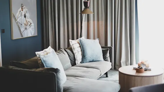 Grå sofa med blå puter står i en moderne stue. Et rundt trebord med stearinlys er i nærheten. Et maleri av ballettdansere henger på den blå veggen.