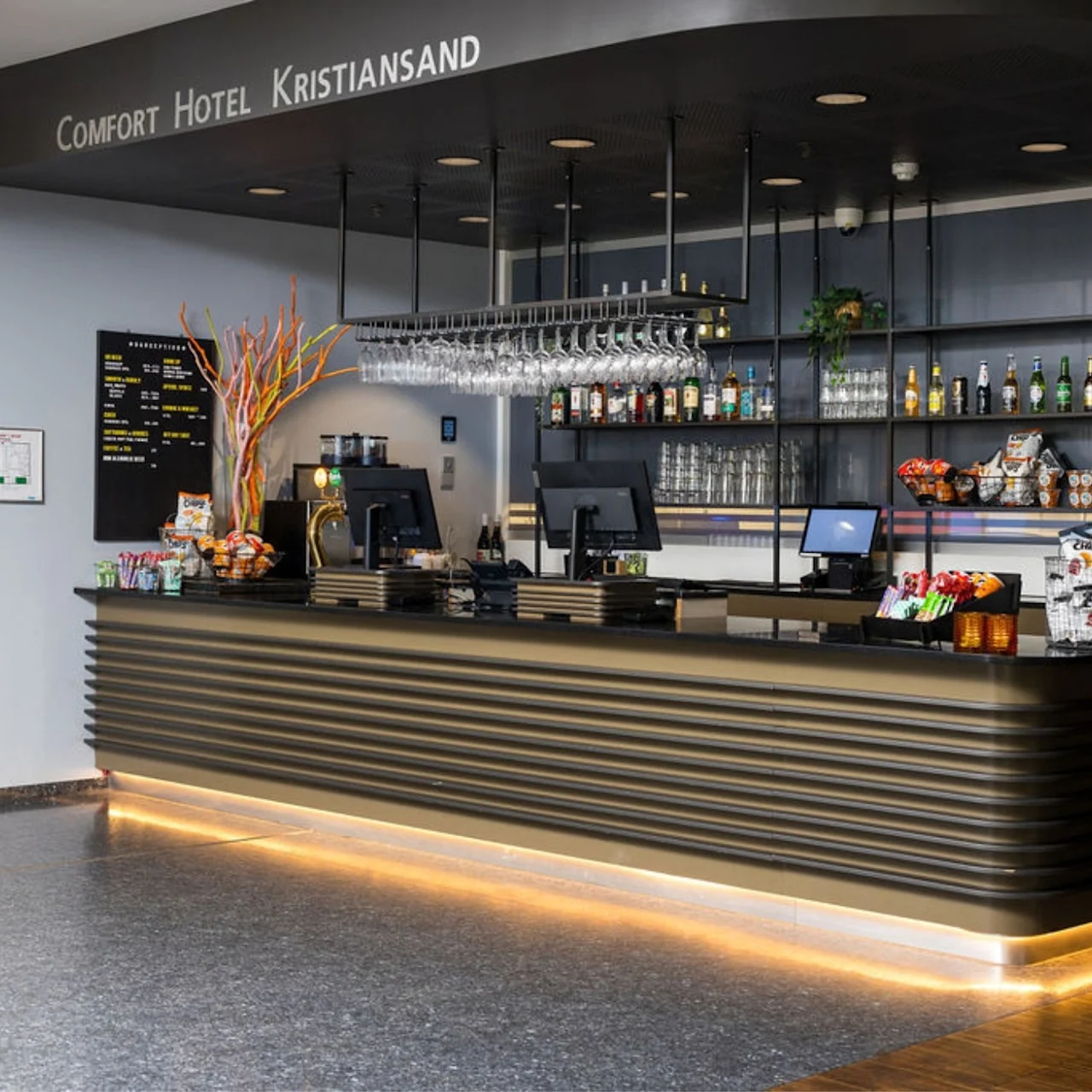 A modern hotel reception desk displays snacks and drinks under hanging glasses in a well-lit lobby. Above, the sign reads "COMFORT HOTEL KRISTIANSAND."