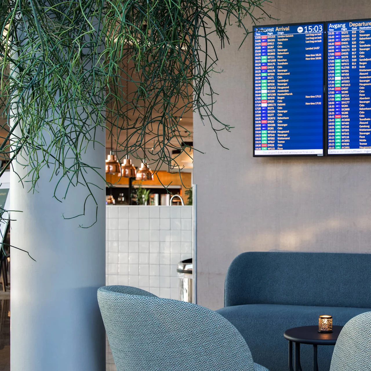 Three digital screens display flight information above a cozy airport lounge with blue seating, surrounded by hanging plants and nearby café with copper light fixtures. 