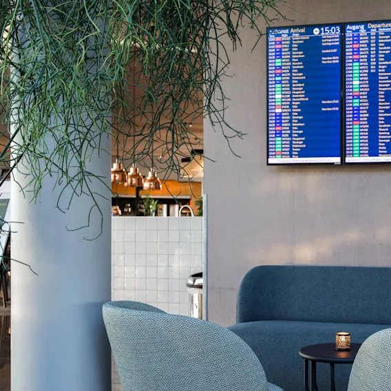 Tre digitale skjermer viser flyginformasjon over en koselig flyplasslounge med blå seter, omgitt av hengende planter og en nærliggende kafé med kobberlamper.