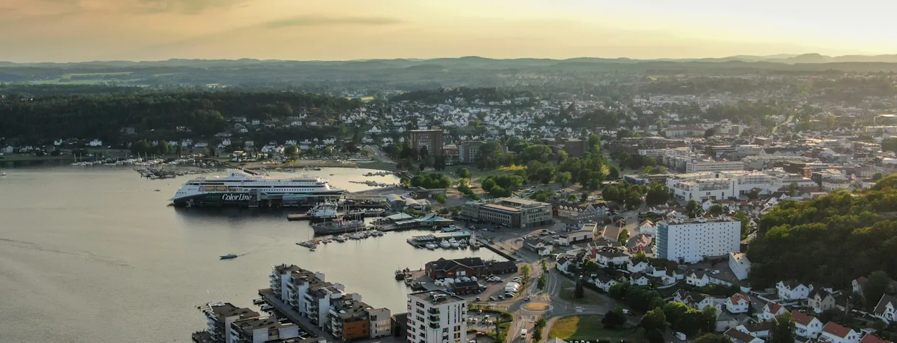 Ilmakuva vilkkaasta rannikkokaupungista auringonlaskun aikaan, suuri lautta satamassa ja rakennuksia rannalla.
