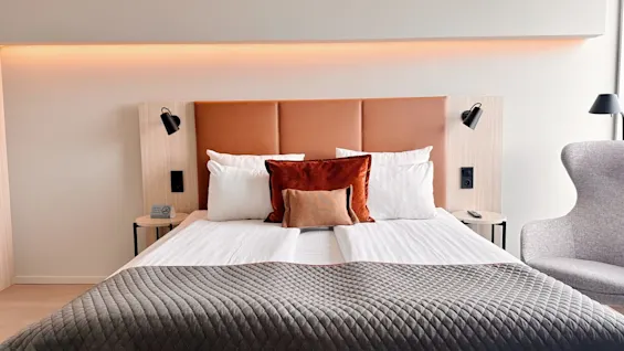 A neatly made bed with brown and white pillows rests in a modern room; a chair and lamps add to the cozy setting. Text: "No smoking."