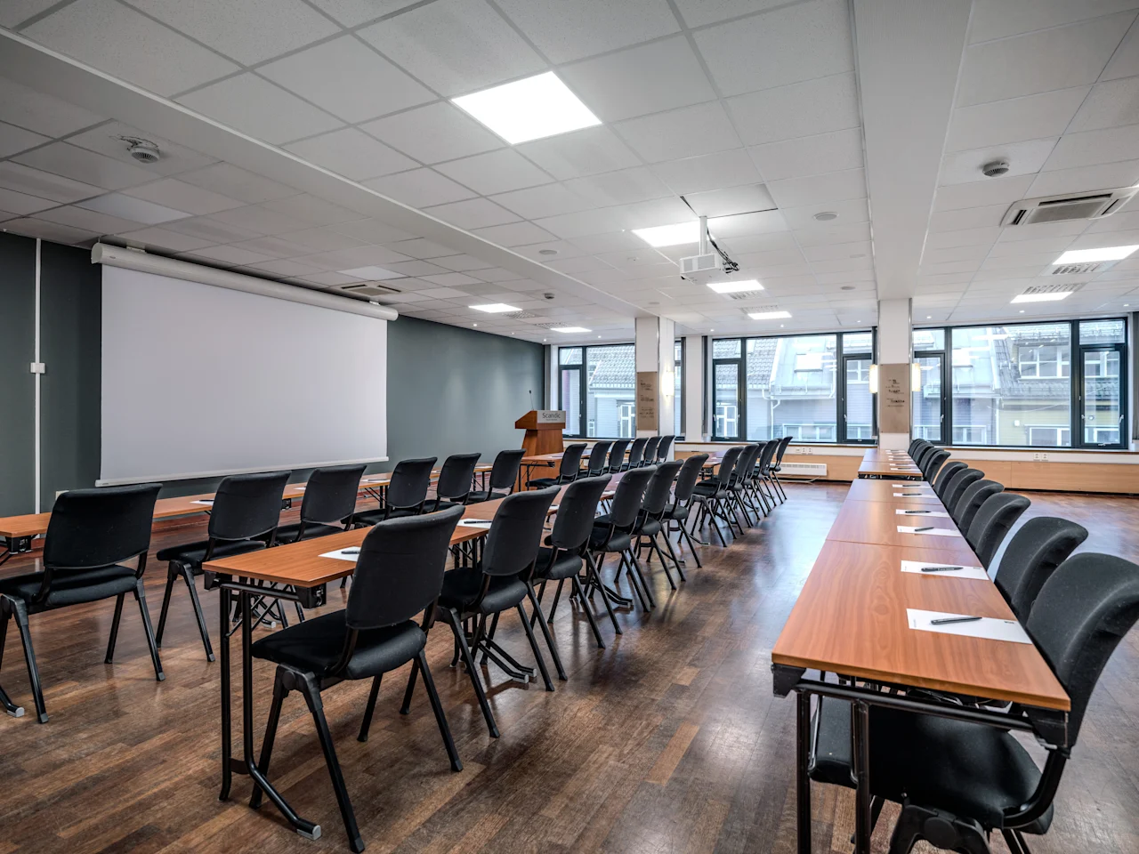 Tilava kokoushuone, jossa on rivejä pöytiä ja tuoleja suurta valkokangasta ja puhujakoroketta kohti, valmiina esitykseen.