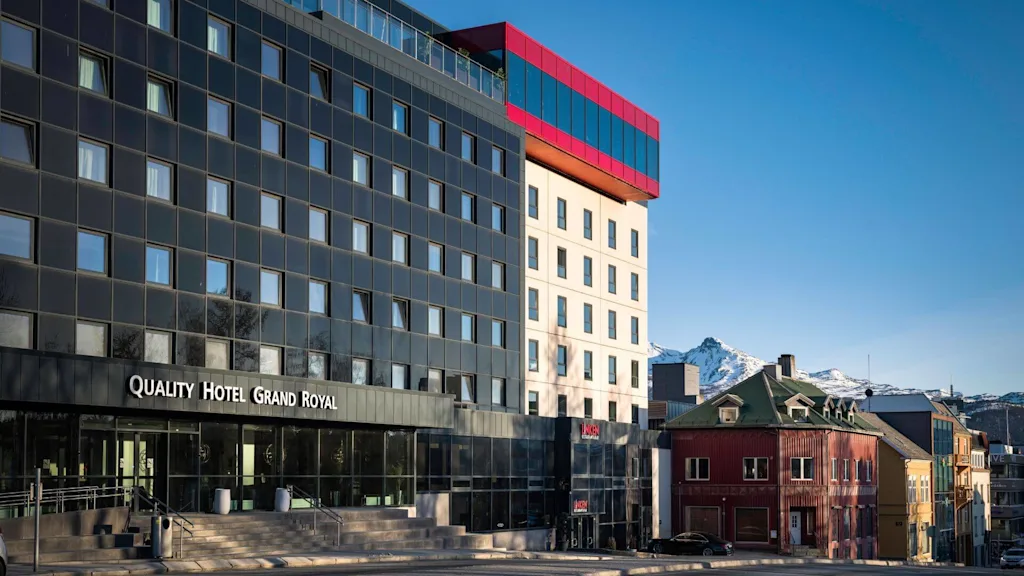En moderne, fleretasjes hotellbygning merket "Quality Hotel Grand Royal" står fremtredende. Ved siden av, kompletterer en rød struktur designen. Snødekte fjell er synlige i bakgrunnen.