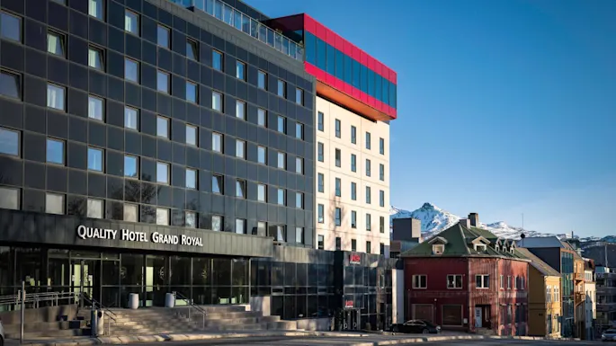A modern, multi-story hotel building labeled "Quality Hotel Grand Royal" stands prominently. Adjacent, a red structure complements the design. Snow-capped mountains are visible in the background.
