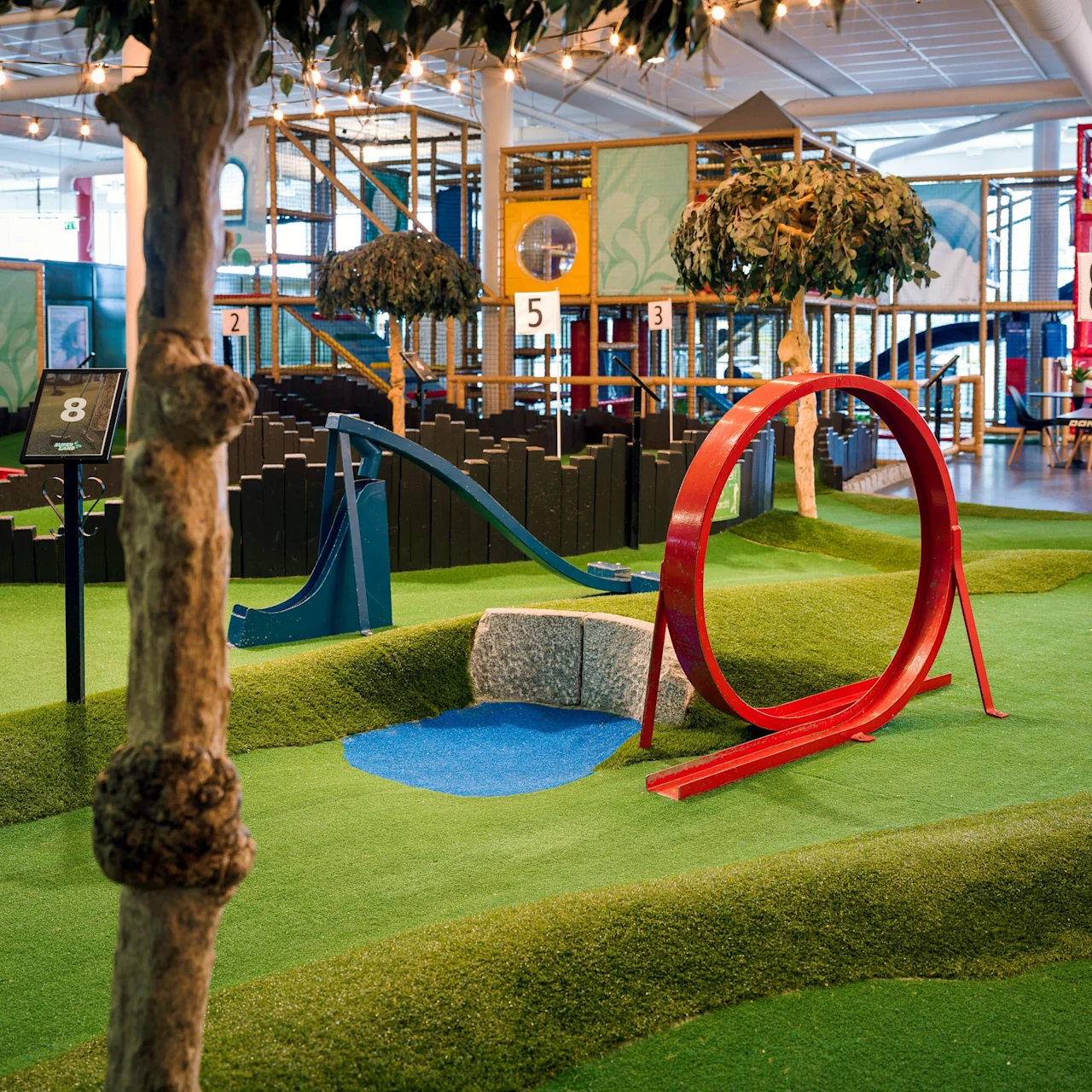 A mini-golf course with a red loop obstacle and a blue ramp sits on artificial grass in an indoor amusement park. Numbered signs and faux trees decorate the scene.