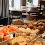 Bread and fruit are arranged on a table in a buffet setting, surrounded by dining tables and chairs in a softly lit room with large windows and green plants.