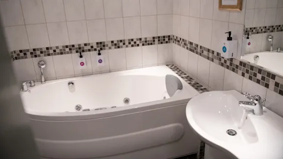 A white bathtub is fitted with two soap dispensers on a tiled wall, next to a sink in a bathroom. A framed sign reads: "Splash, relax, take a bath!"