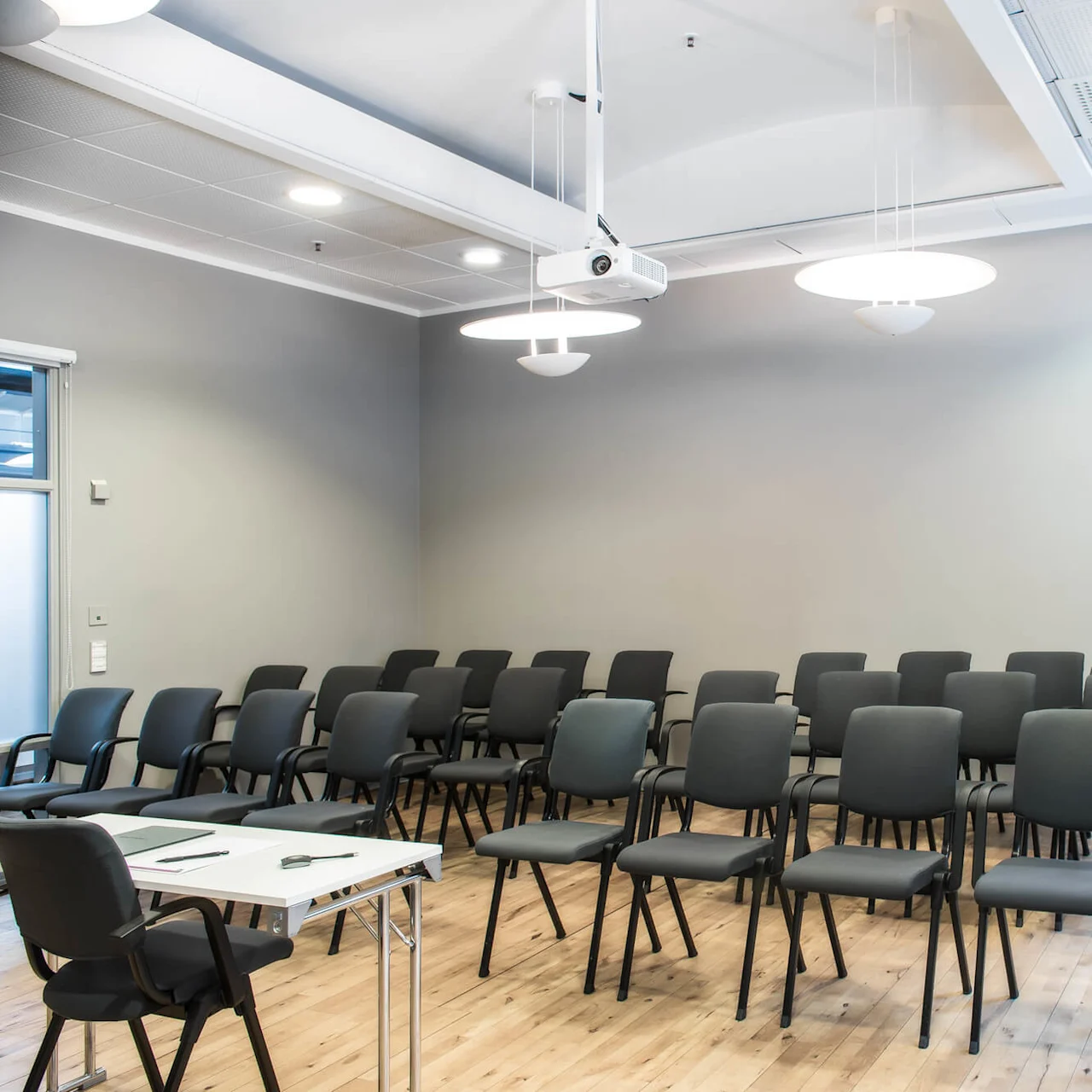 A small conference room with rows of empty black chairs facing a white table, set in a modern space with gray walls, wood flooring, and overhead lights.