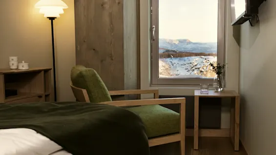 Et hyggeligt værelse med seng, lænestol og et stort vindue med en fantastisk udsigt over sneklædte bjerge.