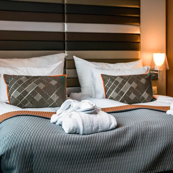 To hvite badekåper ligger fint på en seng med et grått teppe i et varmt opplyst hotellrom med mønstrede puter og en polstret hodegjerde, som gir en koselig atmosfære.